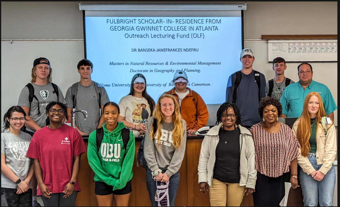15 people standing in the front of a classroom with a presentation projected on the wall behind them. The Fulbright scholar is in the bottom row, third from left.